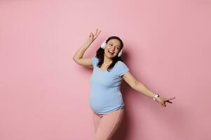 joyeux Latin américain Enceinte femme écoute à la musique dans sans fil écouteurs et dansant plus de rose studio Contexte photo