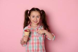 peu enfant fille sourit à caméra, montrant une gaufre cône avec délicieux la glace crème sur isolé rose Contexte photo