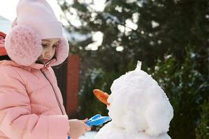 fermer portrait charmant peu enfant fille dans rose vers le bas veste et duveteux cache-oreilles, bâtiment bonhomme de neige dans neigeux arrière-cour photo