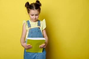 portrait de mignonne enfant fille dans Jaune T-shirt et bleu denim robe salopette, en portant cahier de texte isolé studio Contexte photo
