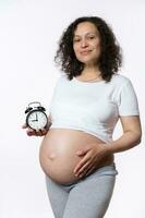 magnifique Enceinte femme coups sa ventre, sourit et spectacles alarme l'horloge avec compte à rebours à le attendu Date de naissance. photo