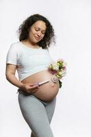 content souriant adulte gravide femme en portant positif grossesse tester et bouquet de fleurs plus de sa gros Enceinte ventre photo