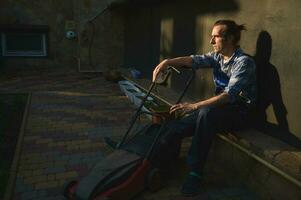 portrait de une fatigué surmené jardinier homme avec électrique pelouse tondeuse, relaxant après difficile travail journée dans le arrière-cour photo