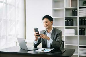 Jeune asiatique affaires homme en utilisant portable et tablette tandis que séance à sa travail lieu. concentré à travail. photo