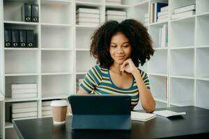 Jeune africain étudiant en train d'étudier à le Université bibliothèque, séance à le bureau, en utilisant une portable ordinateur, tablette et écouteurs ayant une vidéo discuter. photo