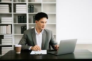 Jeune asiatique affaires homme en utilisant portable et tablette tandis que séance à sa travail lieu. concentré à travail. photo