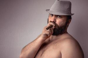 bel homme barbu à moitié nu avec un chapeau gris fumant son cigare et debout contre un mur de fond gris. photo
