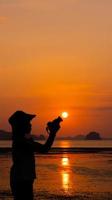 silhouette de jeune femme utilisant un appareil photo pour prendre une photo du coucher de soleil sur la plage pendant les vacances d'été