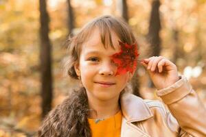 proche portrait de une peu fille. elle couvertures sa œil avec une érable l'automne feuille. tomber saison et les enfants concept. photo