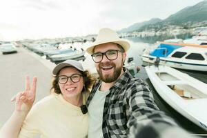 Jeune couple prise une soi portrait en riant comme elles ou ils pose proche ensemble pour caméra sur leur téléphone intelligent en plein air dans été Port Marina avec bateaux et yachts photo