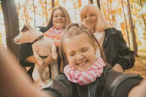 Trois générations de femmes et chien ressentir amusement Regardez à caméra posant pour autoportrait image ensemble, marrant excité enfant, maman et grand-mère avoir amusement prendre plaisir fin de semaine prendre selfie sur gadget dans l'automne photo