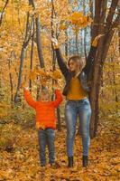 Célibataire parent famille en jouant avec l'automne feuilles dans parc. content maman et fils jeter l'automne feuilles en haut dans tomber parc. photo