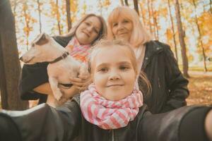 Trois générations de femmes et chien ressentir amusement Regardez à caméra posant pour autoportrait image ensemble, marrant excité enfant, maman et grand-mère avoir amusement prendre plaisir fin de semaine prendre selfie sur gadget dans l'automne photo