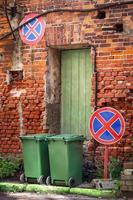deux poubelles debout sur rue avec mur de brique rouge avec porte en bois verte et deux panneaux de signalisation restrictifs photo