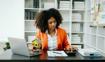 Jeune réel biens agent ouvrier travail avec portable et tablette à table dans Bureau et petit maison à côté de il. photo