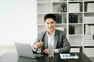 Jeune réel biens agent ouvrier travail avec portable et tablette à table dans moderne Bureau et petit maison à côté de il. photo