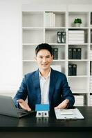 Jeune réel biens agent ouvrier travail avec portable et tablette à table dans moderne Bureau et petit maison à côté de il. photo