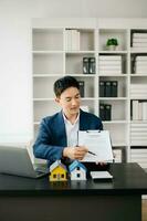 Jeune réel biens agent ouvrier travail avec portable et tablette à table dans moderne Bureau et petit maison à côté de il. photo