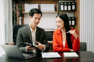 un collègue d'affaires asiatique présente et explique et utilise un ordinateur portable et une tablette. travail d'équipe, équipe de marketing financier, assis dans la salle de bureau. photo