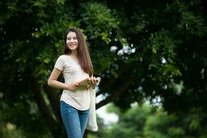 sur de soi souriant Jeune asiatique femme élèves en portant carnet et permanent sur pelouse dans parc. concept de éducation, Campus, adolescent. photo