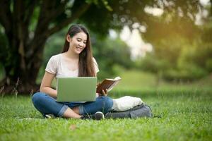 sur de soi souriant Jeune asiatique femme élèves en train de lire et en pensant Faire devoirs avec ordinateur portable sur pelouse dans parc. concept de éducation, Campus, adolescent. photo