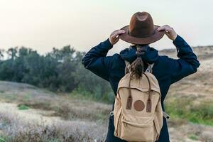 promeneur femme de retour avec bleu veste et sac à dos, détient sa branché chapeau, tandis que à la recherche à le horizon. une randonnée et Voyage concept. photo