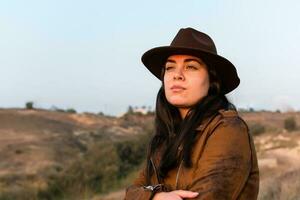 Jeune femme dans chapeau et marron cuir veste, à la recherche dans le distance, dans Naturel parc. photo