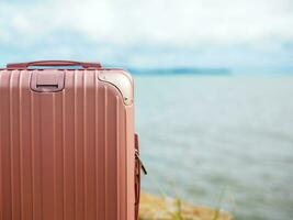 un pastel rose valise posé en dehors dans le Contexte est une flou vue de le mer sur une ensoleillé journée. adapté pour Aller sur vacances photo