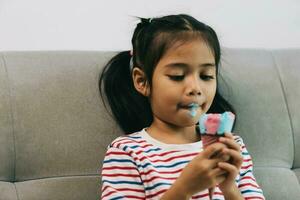 asiatique peu fille en mangeant la glace crème tandis que séance sur le canapé à maison. photo