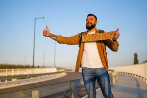 homme est auto-stop sur bord de la route en essayant à Arrêtez auto. il est en portant papier carton avec une inscription. photo