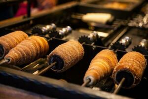 préparation de le traditionnel trdelnik une gentil de cracher gâteau fabriqué de roulé pâte enveloppé autour une bâton, grillé et surmonté avec sucre et noyer mélanger photo