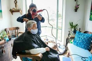 Sénior femme avoir une la Coupe de cheveux à Accueil pendant covid19 pandémie portant visage masque photo