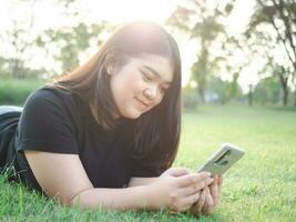 portrait Jeune femme asiatique joufflu mignonne magnifique un la personne porter noir chemise Regardez main en portant en utilisant intelligent téléphone dans jardin parc Extérieur soir lumière du soleil Frais souriant de bonne humeur content se détendre été journée photo