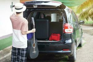 retour vue de asiatique homme détient le guitare et prendre bagage dans le sien voiture.concept, en voyageant, Aller par auto. vacances, vacances ou fin de semaine voyage. préparer et prêt aller. photo