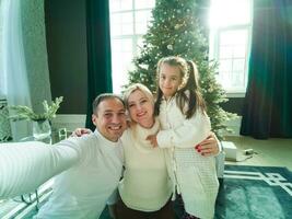 famille, vacances, La technologie et gens - souriant mère, père et peu fille fabrication selfie avec caméra plus de vivant pièce et Noël arbre Contexte photo