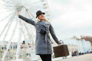 attrayant blond fille dans décontractée vêtements, portant une manteau, en portant une marron rétro valise dans le sien mains, Contexte de le ferris roue photo