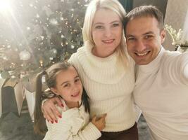 famille, vacances, La technologie et gens - souriant mère, père et peu fille fabrication selfie avec caméra plus de vivant pièce et Noël arbre Contexte photo
