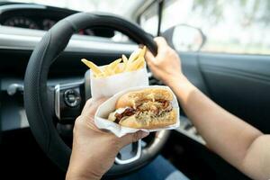 asiatique Dame en portant Hamburger et français frites à manger dans voiture, dangereux et risque un accident. photo