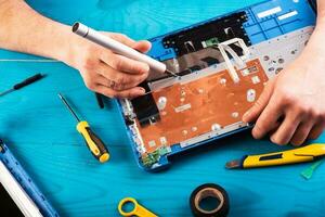 l'assistant répare l'ordinateur portable avec des outils et des mains sur la table en bois bleue. vue de dessus photo