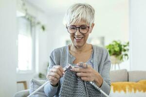 personnes âgées femme dans des lunettes asseoir sur canapé à Accueil sourire en portant tricot aiguilles et fil tricots vêtements pour aimé ceux, préféré activité et Passe-temps, retraité tranquille insouciant la vie concept photo