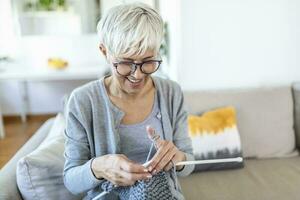 personnes âgées femme dans des lunettes asseoir sur canapé à Accueil sourire en portant tricot aiguilles et fil tricots vêtements pour aimé ceux, préféré activité et Passe-temps, retraité tranquille insouciant la vie concept photo