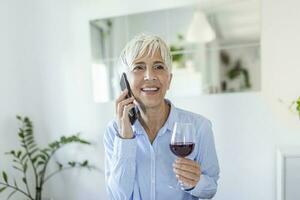 Sénior femme ayant une verre de du vin tandis que en utilisant sa téléphone. content femme relaxant à maison, elle est en buvant une verre de du vin et parlant sur sa téléphone intelligent photo