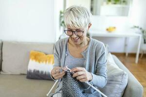 personnes âgées femme dans des lunettes asseoir sur canapé à Accueil sourire en portant tricot aiguilles et fil tricots vêtements pour aimé ceux, préféré activité et Passe-temps, retraité tranquille insouciant la vie concept photo