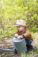 mignonne peu bambin garçon dans une chapeau et caoutchouc bottes est arrosage les plantes avec une arrosage pouvez dans le jardin. une charmant peu enfant portion le sien Parents grandir des légumes. photo