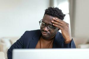 stressé fatigué africain américain homme émouvant temples, Souffrance de mal de crâne après longue heures travail, surmené submergé homme d'affaire séance à bureau, sentiment indisposé photo