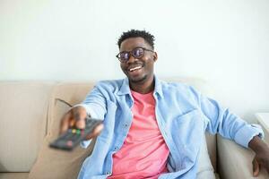 détendu noir gars commutation canaux sur la télé à maison, en portant la télé télécommande, souriant Jeune africain homme séance sur canapé dans vivant chambre, en train de regarder télévision, profiter paresseux fin de semaine photo