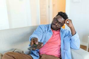 Beau africain américain Jeune homme séance sur le canapé à maison, en train de regarder télévision, relaxant et à la recherche confus photo