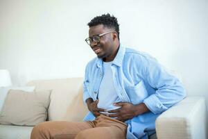 Jeune africain homme en portant le sien estomac dans douleur, homme avec reflux maladie en portant le sien ventre avec douloureux expression photo