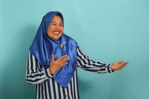 une âge moyen asiatique femme, portant une bleu hijab et une rayé chemise, étend sa mains à le côté, attrayant Quelqu'un à venir, tandis que permanent contre une bleu Contexte photo