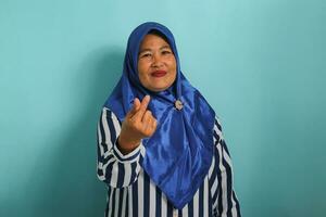 une âge moyen asiatique femme, dans une bleu hijab et rayé chemise, fait du une mini cœur main signe ou coréen doigt cœur signe, isolé sur une bleu Contexte. photo